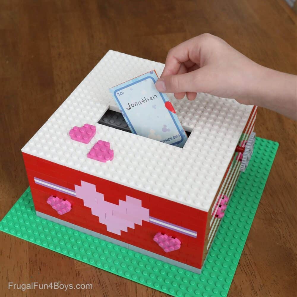A cool LEGO Valentine box idea featuring a child's hand putting a paper Valentine card into the opening on top of a box made out of white, red, and pink LEGO bricks with heart patterns on it.