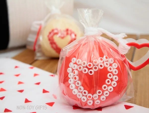 Two DIY Valentines consisting of red and white homemade playdough with pony beads positioned in a heart on it and then packaged inside a clear treat bag with a heart-shaped cookie cutter tied on.