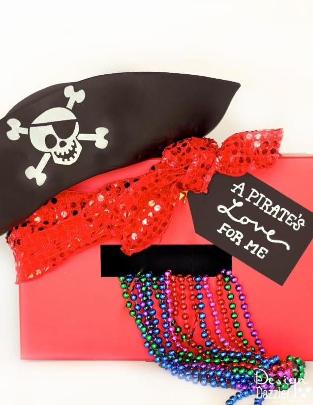 An easy pirate Valentine box idea featuring a red cardboard box positioned on its side with the open slot facing the viewer and shiny metallic party beads spilling out, a red sequined scarf and a kids black pirate costume hat with a white skull on it positioned on the upper left corner, and a black paper tag reading "a pirate's love for me" glued onto the upper right corner.