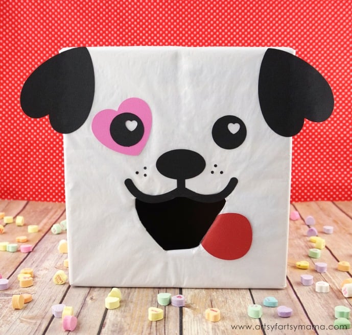 An easy puppy dog Valentine box idea using a white paper-covered box with Cricut-cut cardstock pieces glued, including black upside-down heart ears on each upper corner, black round eyes with a pink heart around one of them, and a black nose and smiling open mouth piece with a red tongue hanging out.