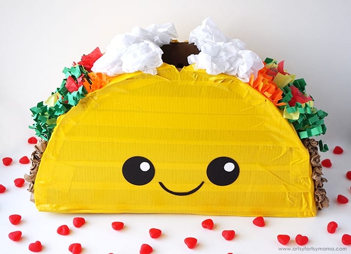 A cute DIY taco Valentine box idea featuring a taco-shaped box made out of painted cardboard with a cute smiley face on it and crumpled tissue paper taco toppings along the top edge.