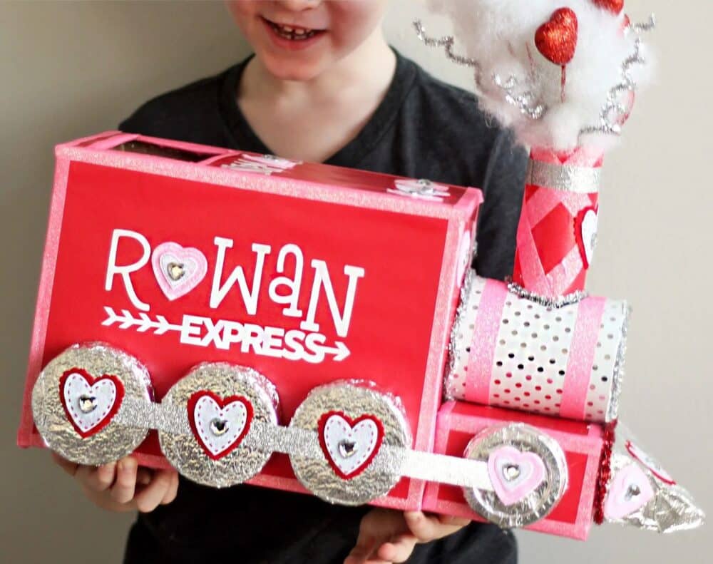 A smiling child holding a creative Valentine box made to look like a train using recycled materials, cardboard boxes, tin foil, red and pink patterned paper, and sparkly silver pipe cleaner and white cotton ball smoke coming out of the cardboard roll smokestack.