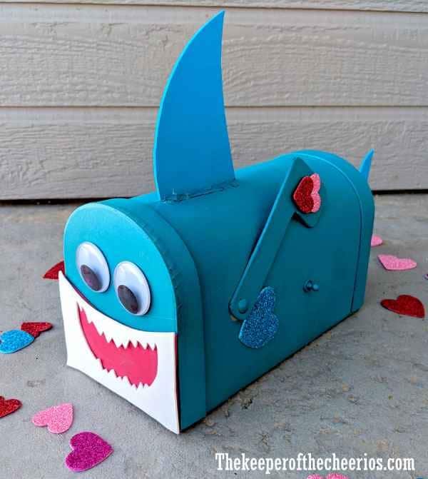 A cute Baby Shark Valentine mailbox craft using a mini craft paper mailbox painted light blue with blue craft foam fins attached to the top and a craft foam tail glued to the back along with white and red craft foam details on the front to look like the shark's mouth and large googly eyes glued on the front.