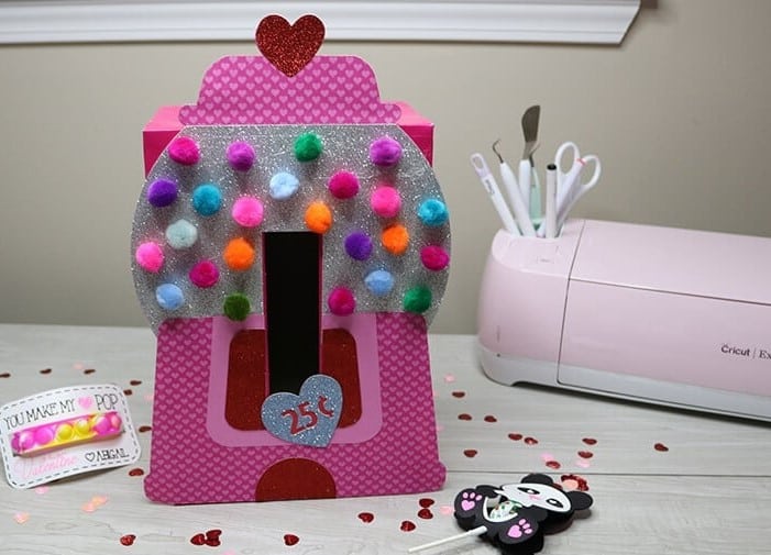 A cute Valentine mailbox craft using a free SVG file for a Cricut cutting machine to cut out cardstock pieces to make a pink, red, and silver gumball machine-shaped Valentine card mailbox with the slot opening cut out of the front to look like the coin slot and pom pom gumballs glued onto it.