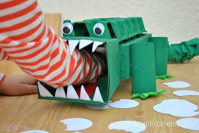 A cool alligator Valentine box idea for kids featuring a child's arm reaching into the open mouth of a DIY Valentine's box crafted to look like an alligator using painted cardboard boxes, cardboard egg cartons, toilet paper rolls, and googly eyes.