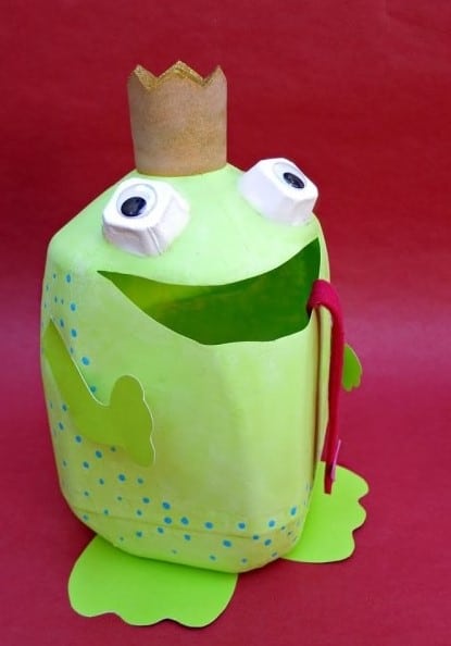 A creative Valentine box idea using a gallon-sized plastic milk jug painted lime green with a wide mouth opening cut out near the top, white painted egg carton cup eyes with black circle pupils, a gold painted paper crown on top, a long red felt tongue hanging out of the mouth, and lime green paper arms and feet glued on.